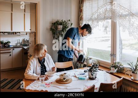 Pflegender Mann wäscht Pflanzen durch Seniorin beim Essen, während sie zu Hause am Esstisch sitzt Stockfoto