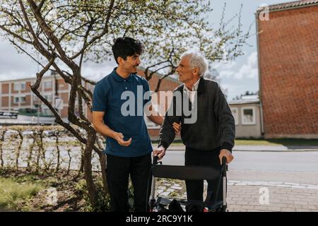 Lächelnder Pfleger, der Seniorenhelfer mit Gehhilfe auf dem Fußweg unterstützt Stockfoto