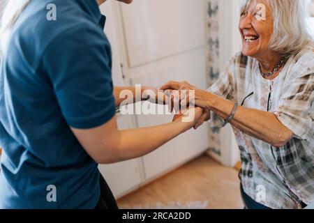 Glückliche Seniorin, die Händchen hält, während sie zu Hause mit einer Pflegekraft spricht Stockfoto