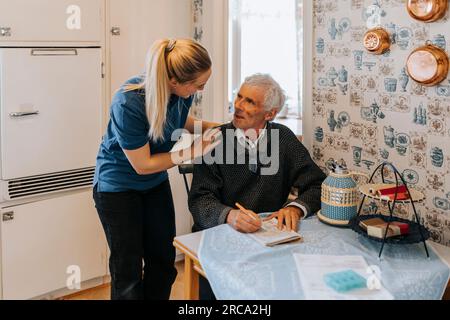 Weibliche Pflegehelferin, die mit einem Seniorenmann spricht, der zu Hause Soduko macht Stockfoto
