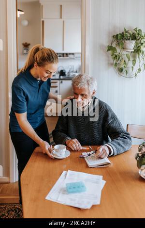 Pflegehelferin serviert Tee an Seniorenmann, der zu Hause sitzt Stockfoto