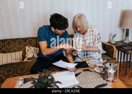 Pflegender Mann mit medizinischen Berichten, die Seniorin erklären, die zu Hause Smartphone benutzt Stockfoto
