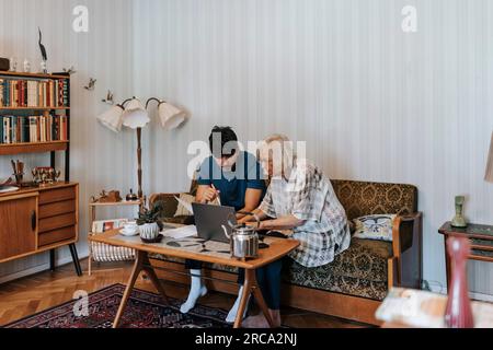 Seniorin mit Laptop und Pflegehelferin, die zu Hause auf dem Sofa sitzt Stockfoto