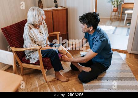 Pflegehelferin, die Seniorin hilft, während sie zu Hause Socken trägt Stockfoto