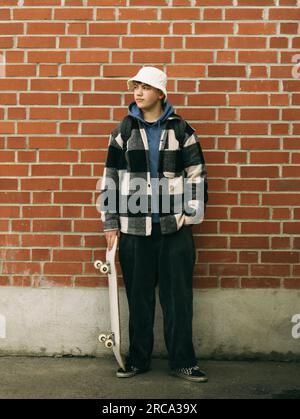 Junger Mann mit Skateboard, der vor einer Ziegelwand posiert Stockfoto