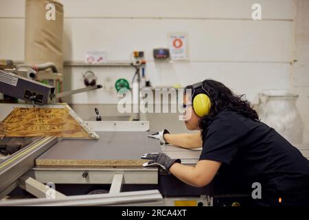 Seitenansicht einer Zimmerlerin mit Schutzbrille und Gehörschutz bei der Arbeit im Werk Stockfoto