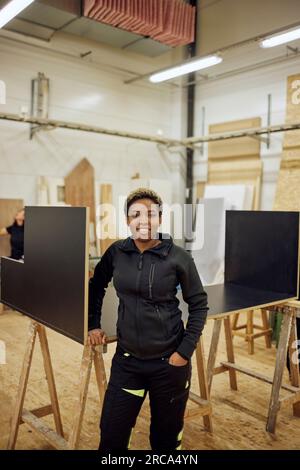 Porträt einer lächelnden jungen Zimmerlerin, die in der Werkstatt mit der Hand in der Tasche steht Stockfoto