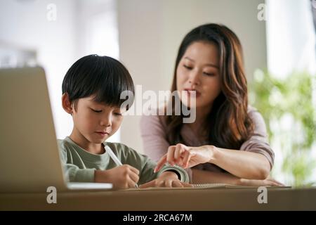 Junge asiatische Mutter, die zu Hause am Tisch sitzt und dem Sohn beim Lernen hilft Stockfoto