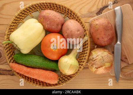 Rohes Gemüse, rohe Kartoffeln, süßer Pfeffer, Zwiebeln, Karotten, Tomate und Gurke mit Tonplatte und Metallmesser auf Holztisch, Draufsicht. Stockfoto