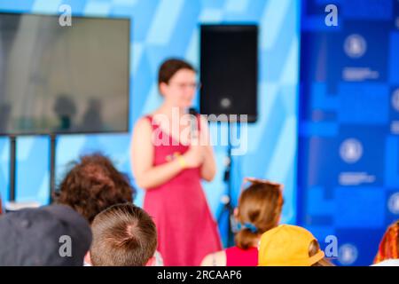 Rückansicht von nicht wiedererkennbaren Personen, die auf Stühlen sitzen und anonymen weiblichen Sprechern auf der Bühne lauschen, während während b das Mikrofon in der Nähe des Monitors steht Stockfoto