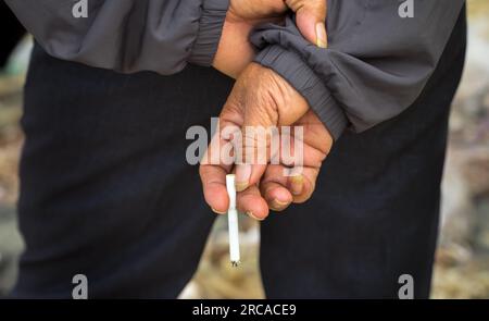 Nahaufnahme eines vietnamesischen Mannes mit langen Fingernägeln, der eine frisch beleuchtete Zigarette hinter seinem Rücken in Danang, Vietnam, hält. Stockfoto