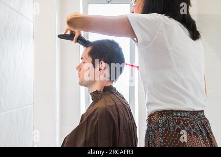 Eine Frau, die einem weißen Mann einen Haarschnitt in einem Heim verpasst. Familienleben und sorgenfreier moderner Lebensstil. Konzept von Komplizenschaft und Vertrauen. Schließen Stockfoto