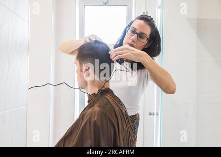 Eine Frau, die einem weißen Mann einen Haarschnitt in einem Heim verpasst. Familienleben und sorgenfreier moderner Lebensstil. Konzept von Komplizenschaft und Vertrauen. Schließen Stockfoto