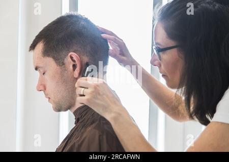 Eine Frau, die einem weißen Mann einen Haarschnitt in einem Heim verpasst. Familienleben und sorgenfreier moderner Lebensstil. Konzept von Komplizenschaft und Vertrauen. Schließen Stockfoto