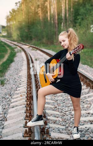 Ein 13-jähriger Teenager mit einer Gitarre. Das Mädchen reist mit einer Gitarre. Das Mädchen steht auf den Schienen, geht in die Ferne, im Wald bei Sonnen Stockfoto