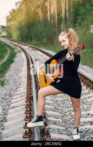 Ein 13-jähriger Teenager mit einer Gitarre. Das Mädchen reist mit einer Gitarre. Das Mädchen steht auf den Schienen, geht in die Ferne, im Wald bei Sonnen Stockfoto