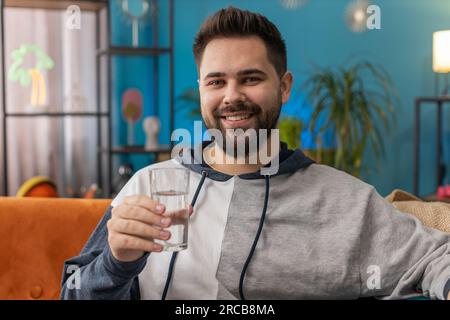 Durstiger Mann hält ein Glas natürliches Aqua trinken Schluck stilles Wasser verhindern Austrocknung sitzt zu Hause im Wohnzimmer. Weißer Typ mit guten Lebensgewohnheiten, gesundes Schlanken, Gewichtsverlust Konzept Stockfoto
