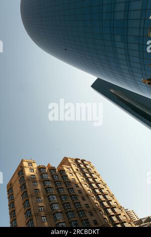 Baku, Aserbaidschan - 26. Juni 2023: Die spiegelnde Oberfläche eines zylindrischen Wolkenkratzers spiegelt ein historisches Gebäude in Baku unter einem klaren Morgenhimmel wider Stockfoto