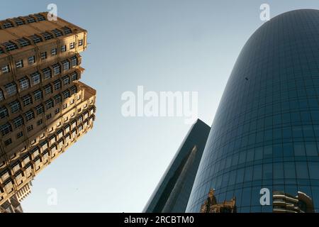 Baku, Aserbaidschan - 26. Juni 2023: Die spiegelnde Oberfläche eines zylindrischen Wolkenkratzers spiegelt ein historisches Gebäude in Baku unter einem klaren Morgenhimmel wider Stockfoto