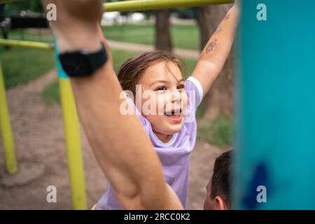 Kleine Tochter spielt mit Vater beim Workout im Fitnessraum im Freien. Fröhliche Gefühle. Gesunder Lebensstil in der Familie Stockfoto