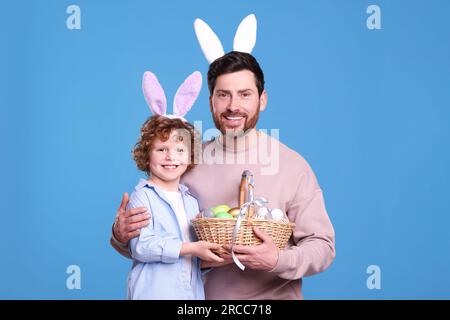 Glücklicher Vater und Sohn in süßen Hasenohren Stirnbänder halten Osterkorb mit bemalten Eiern auf hellblauem Hintergrund Stockfoto