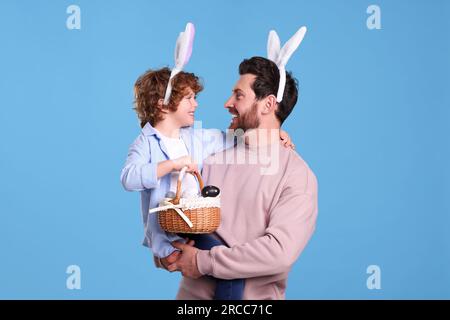 Glücklicher Vater und Sohn in süßen Häschenohren Stirnbänder auf hellblauem Hintergrund. Ein Junge mit einem Osterkorb und bemalten Eiern Stockfoto