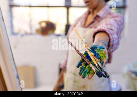 Mittelteil einer birassischen Frau, die Pinsel mit Farbe bedeckt im Studio, Kopierraum Stockfoto