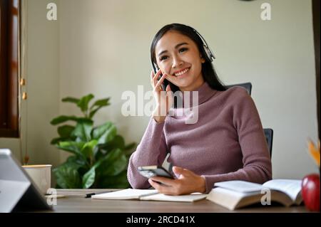 Eine attraktive und glückliche Asiatin hört gerne Musik über ihre kabellosen Kopfhörer an ihrem Schreibtisch. Stockfoto