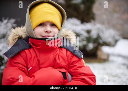 Nahaufnahme eines gut aussehenden, kaukasischen Teenagers im Alter von 11-13 Jahren, in warmer roter Daunenjacke und gelbem Wollhut, lächelnd in die Kamera, stehend Stockfoto