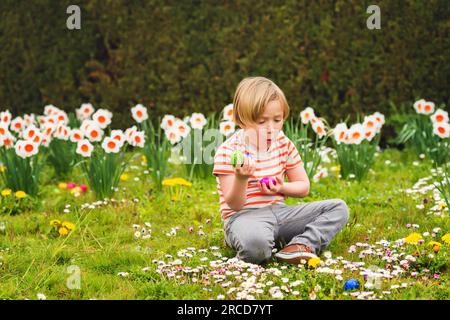 Ein bezaubernder kleiner blonder Junge, der im Park mit bunten ostereiern spielt, Eierjagd Stockfoto