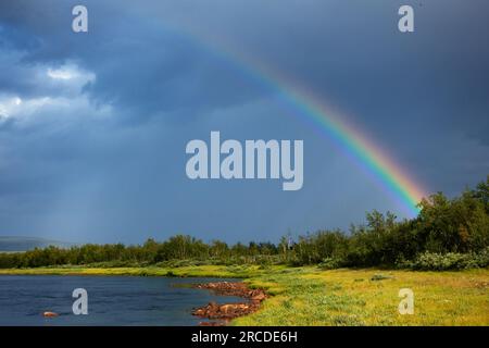 Regenbogen am Flussufer des Lainio im schwedischen Lappland mit dramatischem dunklen Himmel und Wolken im Hintergrund am Sommernachmittag. Stockfoto