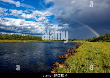 Regenbogen am Flussufer des Lainio im schwedischen Lappland mit dramatischem dunklen Himmel und Wolken im Hintergrund am Sommernachmittag. Stockfoto