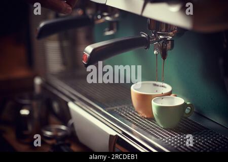 Selektiver Fokus des Espressomaschers, der den Kaffeestrahl in zwei Tassen gießt. Orange und Grün. Stockfoto