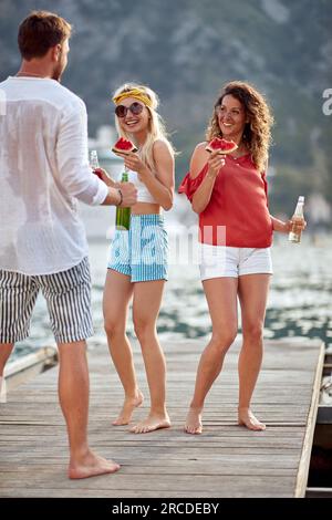 Junge Frauen tanzen mit Bier und einem Stück Wassermelone mit einem Mann auf einem Holzsteg auf dem Wasser. Spaß, Zusammensein, Reisekonzept. Stockfoto