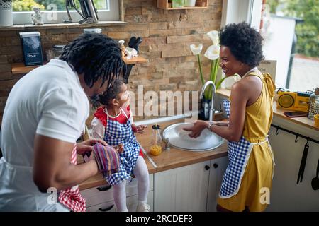 Familienharmonie, die Mutter wäscht sorgfältig das Geschirr in der Küche, während der Vater es sorgfältig trocknet. Ihr kleines Mädchen sitzt in der Küche Stockfoto