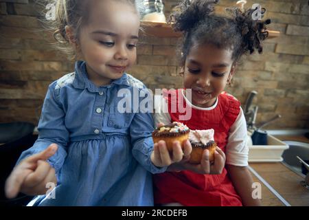 Nahaufnahme des Porträts von zwei kleinen Mädchen, die Muffins in der Küche essen und sich glücklich fühlen. Zuhause, Familie, Lifestyle-Konzept. Stockfoto
