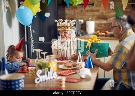 Fröhliche Seniorin, die ein Geburtstagsgeschenk von ihrem Ehemann erhält, mit ihrer Enkelin am Küchentisch sitzt, mit Baloons und Ornamenten im Hinterhof. Fa Stockfoto