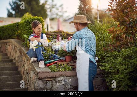 Vater und seine Tochter sind sorgfältig geschnittene Blumen. Das Sonnenlicht badet den Garten und strahlt ein warmes Licht auf ihre Gesichter, während sie zusammenarbeiten. Stockfoto