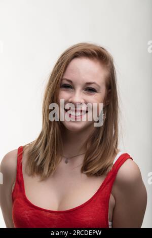 Hellhäutige, erdbeerblonde Frau in einem auffälligen roten Kleid. Ihr lächelnder, lachender Ausdruck trifft auf die Blicke anderer in ihrem halblangen Porträt. Ein reichlicher Bulle Stockfoto