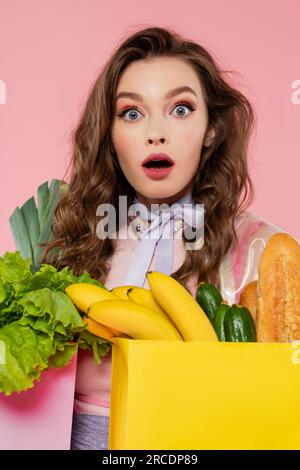 Hausfrauenkonzept, schockierte Frau, die Einkaufstüten mit Gemüse und Bananen trägt, Modell mit welligem Haar auf pinkfarbenem Hintergrund, Konzeptfotografie Stockfoto