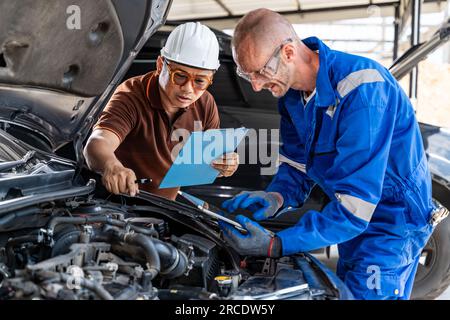 Ein asiatischer Mechaniker-Supervisor, der dem männlichen Automechaniker Reparaturvorschläge gibt, der sich auf seinem Tablet Notizen über den Motor macht Stockfoto