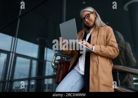 Erwachsene Freiberuflerin mit grauen Haaren in Brillen, die mit einem Laptop vor dem Hintergrund eines Business Centers auf der Straße stand Stockfoto