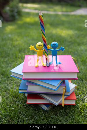 Ein Stapel bunter Bücher liegt auf dem Gras, auf dem zwei menschliche Souvenirfiguren mit einem großen Bleistift stehen. Postkarte für Lehrer. Zurück in die Schule. Konzept o Stockfoto