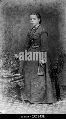 Ein langes Porträt einer jungen Dame in einem langen Kleid, deren Hand auf einem Stuhl ruht, der mehrere Bücher mit Lederbindung enthält. Originalfotograf unbekannt. Aus einer Sammlung viktorianischer Carte de Visite Fotografien aus den 1890er Jahren. Carte de visite ist ein französischer Begriff, der auf Englisch als „Visitenkarte“ bezeichnet wird. Normalerweise wurde ein Fotodruck aus Albumen von etwa 54 x 89 mm auf einer etwas größeren Karte angebracht. In einigen Fällen haben die Verwüstungen der Zeit ihren Tribut gefordert. Stockfoto