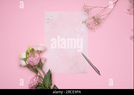 Zwei luxuriöse Edelmetallringe, ein silberner Stift und eine Hochzeitseinladung für die frisch Vermählten auf pinkfarbenem Hintergrund mit wunderschönen Blumen. Das Konzept o Stockfoto