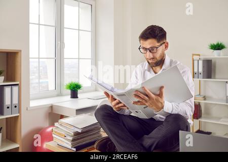 Ein komischer, schockierter, verwirrter Mann, der auf seinem Schreibtisch sitzt und sich einen offenen Papierordner ansieht Stockfoto