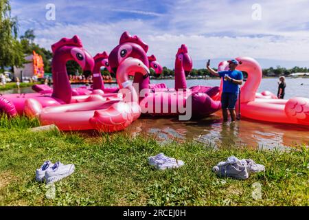 Freiburg, Deutschland. 14. Juli 2023. Drei Paar Schuhe stehen am Ufer des Lake Tunis, während ein Mann im Hintergrund vor schwimmenden, aufblasbaren Flamingos steht und ein Selfie macht. Mit mehr als 150 internationalen Musikern auf sieben Bühnen und mehreren Zehntausenden Besuchern ist es eines der größten Festivals für elektronische Musik im Südwesten. Mit dem Schlagwort „Beach Republic“ wird das Festival für seine besondere Lage, einschließlich des Badestrands am See, werben. Kredit: Philipp von Ditfurth/dpa/Alamy Live News Stockfoto