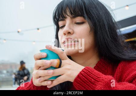 Eine junge, wunderschöne lateinisch-venezolanische Frau in einem roten Pullover sitzt draußen und trinkt an einem bewölkten und kalten Tag ein heißes Getränk, Menschenkonzept, Kopierraum. Stockfoto