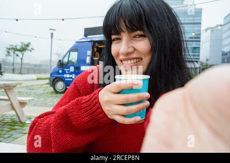 Selfie-Porträt einer jungen, glücklichen venezolanischen latina, die draußen sitzt und morgens mit einer Tasse heißem Kaffee am Wochenende lächelt. Stockfoto