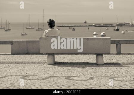 Eine Dame, die allein auf einer Bank sitzt. Sie blickt auf die Cascais Bay und Marina. Cascais, Portugal. Stockfoto